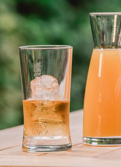 Ein Glas mit einem Eiswürfel und einem goldenen Getränk steht neben einer Karaffe mit orangefarbener Flüssigkeit.