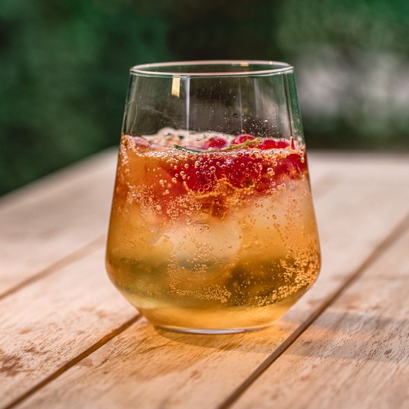Ein Glas mit sprudelndem Getränk, gefüllt mit Eis und frischen Himbeeren, steht auf einem Holztisch im Freien.