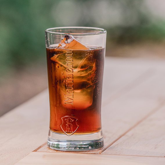 Ein Glas mit einem dunklen Getränk, gefüllt mit Eiswürfeln, steht auf einem Holztisch im Freien.