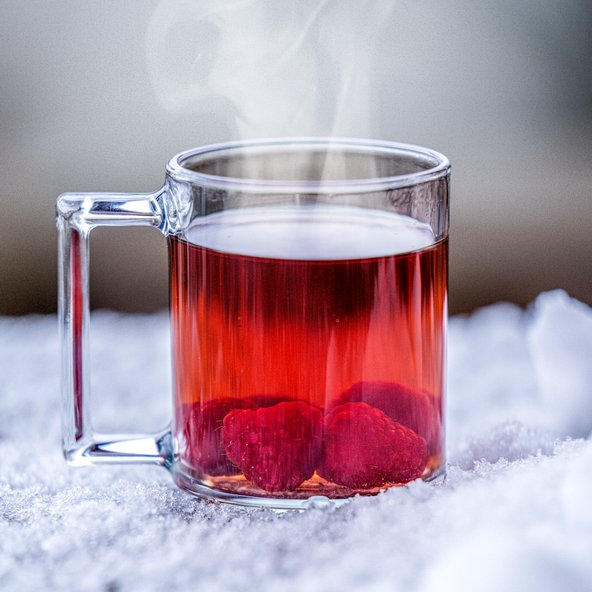 In einem klaren Glas dampft ein fruchtiges Getränk mit Himbeeren, das auf einer kalten, schneebedeckten Fläche platziert ist.
