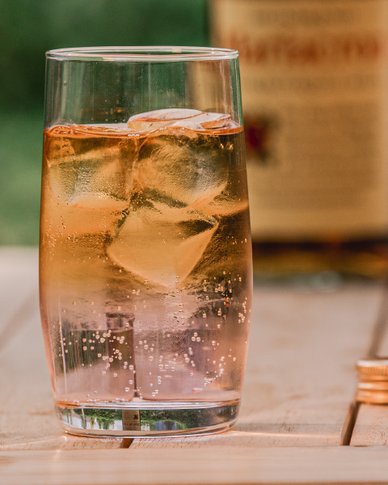 Ein Glas mit sprudelndem Getränk und Eiswürfeln steht auf einem Holztisch, im Hintergrund eine Flasche mit Etikett.