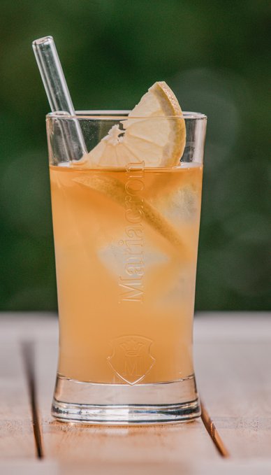 Ein erfrischendes Getränk in einem Glas mit einer Zitronenscheibe und einem Strohhalm, im Hintergrund eine Flasche Likör.
