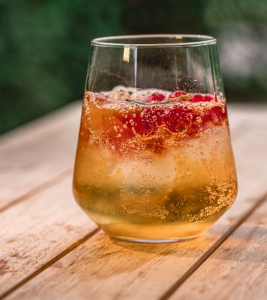 Ein Glas mit sprudelndem Getränk, gefüllt mit Eis und frischen Himbeeren, steht auf einem Holztisch im Freien.