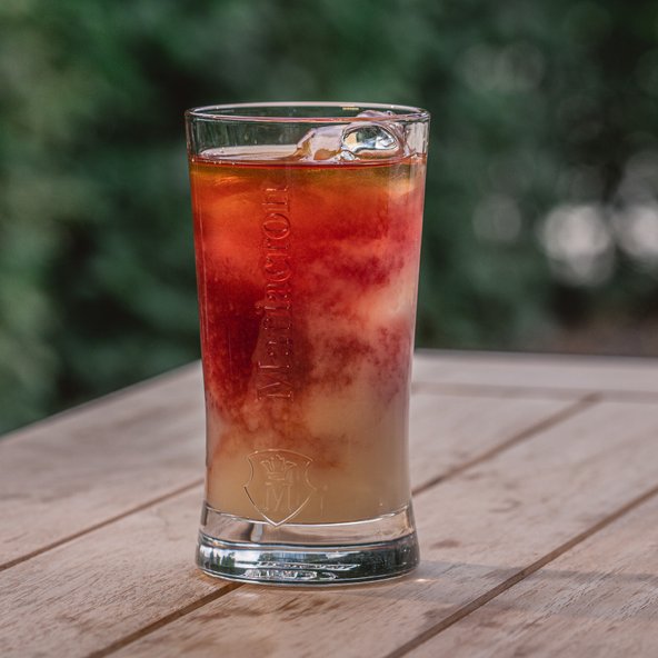 Ein Glas mit einem bunten Getränk, das Eiswürfel enthält, steht auf einem Holztisch vor einem unscharfen grünen Hintergrund.