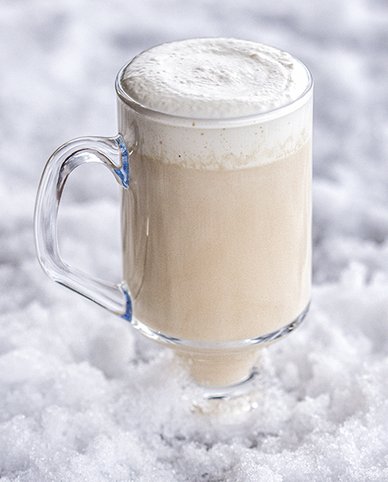 Ein Glas mit schaumigem Getränk steht auf einer schneebedeckten Fläche, das Getränk hat eine cremige, beige Farbe.