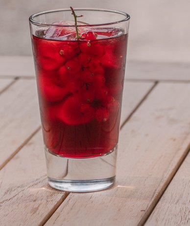 Ein Glas mit einem erfrischenden, roten Getränk, gefüllt mit Eis und frischen roten Johannisbeeren, steht auf einem Holztisch.
