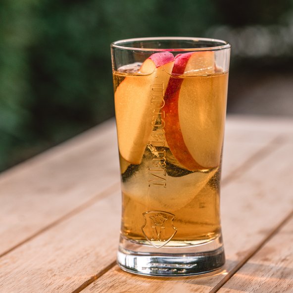 Ein Glas mit einem erfrischenden Getränk, gefüllt mit goldener Flüssigkeit und Apfelscheiben, steht auf einem Holztisch.