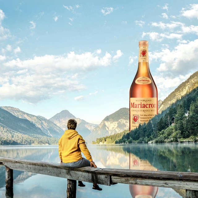 Ein ruhiger Moment am See, wo ein Mann auf einem Holzsteg sitzt und die beeindruckende Natur mit einer großen Flasche Mariacron betrachtet.