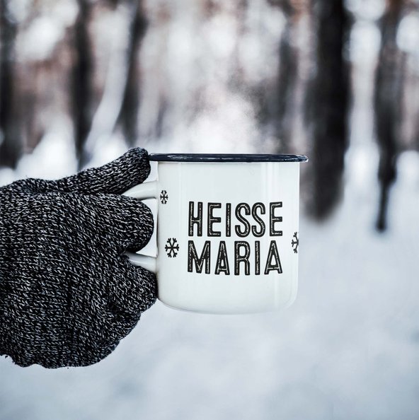 Eine Hand in einem grauen Handschuh hält eine dampfende Tasse mit der Aufschrift 'HEISSE MARIA' vor einer verschneiten Waldlandschaft.