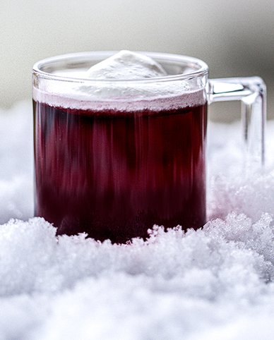 Ein Glas mit einem leuchtend roten Getränk, das auf einer weichen Schneedecke platziert ist, verleiht eine winterliche Atmosphäre.