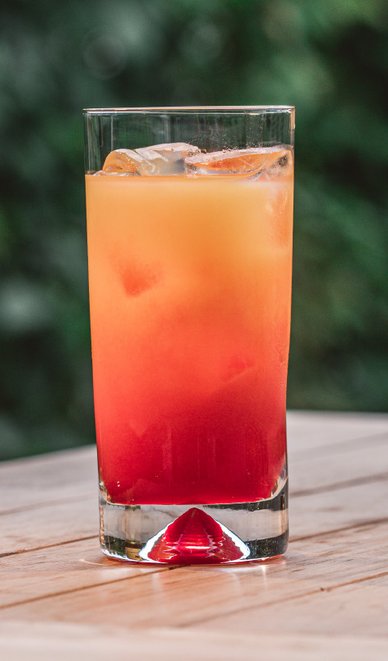 Ein erfrischendes Getränk in einem hohen Glas, mit Eiswürfeln und einer rot-gelben Farbverlauf, auf einem Holztisch.