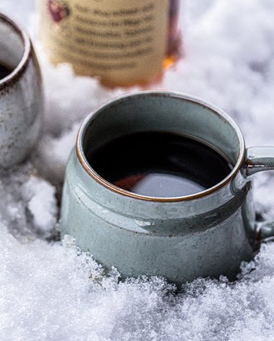 Zwei Tassen mit dampfendem Kaffee stehen in frischem, weißem Schnee, während eine Flasche im Hintergrund sichtbar ist.