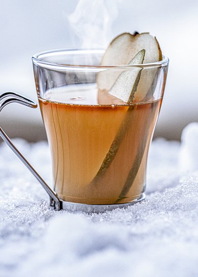 Ein dampfender Tee in einem klaren Glas, garniert mit einer Scheibe Birne, steht auf frischem, weißem Schnee.