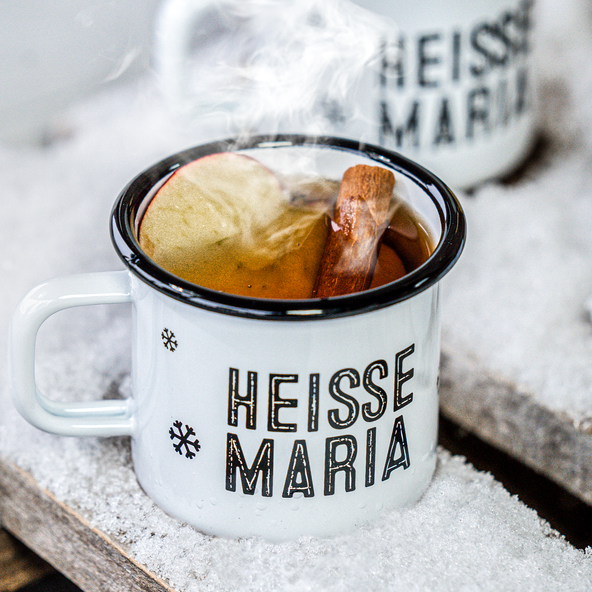 Der Becher 'HEISSE MARIA' zeigt ein aromatisches Getränk mit Zimtstange und Apfel, umgeben von einer schneebedeckten Umgebung.
