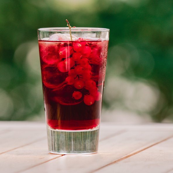 Ein Glas mit einem erfrischenden, roten Getränk, gefüllt mit Eis und frischen roten Johannisbeeren, steht auf einem Holztisch.