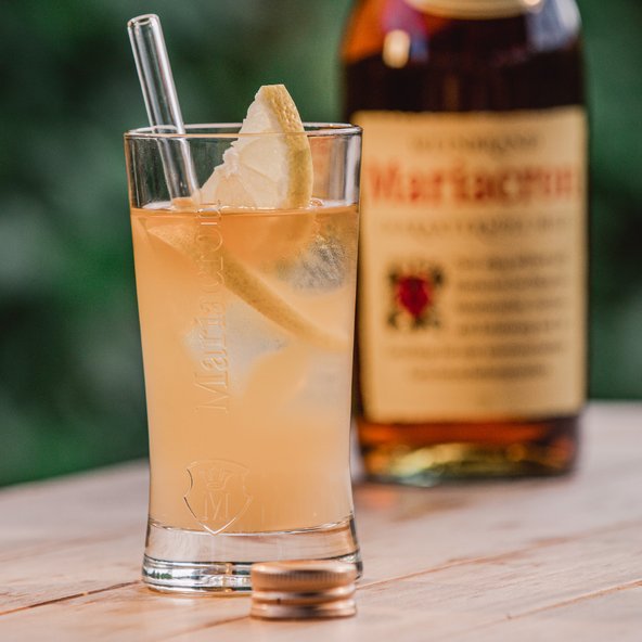 Ein erfrischendes Getränk in einem Glas mit einer Zitronenscheibe und einem Strohhalm, im Hintergrund eine Flasche Likör.