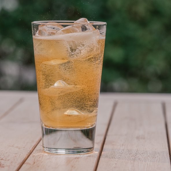 Ein Glas mit einem sprudelnden, goldenen Getränk und Eiswürfeln steht auf einem Holztisch im Freien.