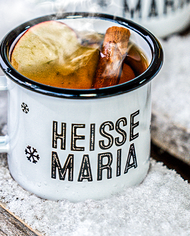 Ein dampfender Becher mit dem Aufdruck 'HEISSE MARIA', gefüllt mit einem warmen Getränk und Zimtstange, steht auf Schnee.