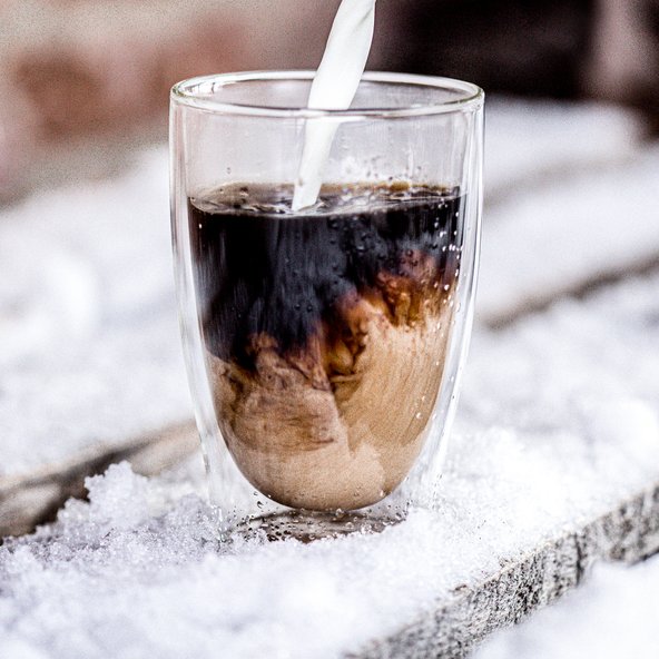 Ein Glas mit Kaffee, in das frische Milch gegossen wird, während es auf einer schneebedeckten Holzoberfläche steht.