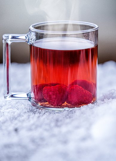 In einem klaren Glas dampft ein fruchtiges Getränk mit Himbeeren, das auf einer kalten, schneebedeckten Fläche platziert ist.