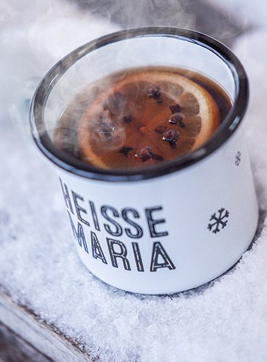 Ein dampfender Becher mit heißem Getränk, garniert mit einer Orangenscheibe und Gewürznelken, steht auf schneebedecktem Untergrund.