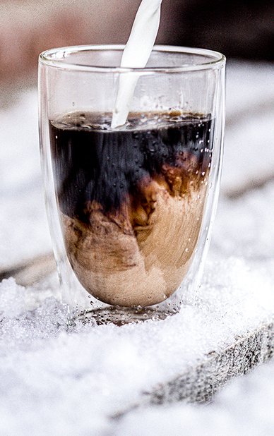 Ein Glas mit Kaffee, in das frische Milch gegossen wird, während es auf einer schneebedeckten Holzoberfläche steht.