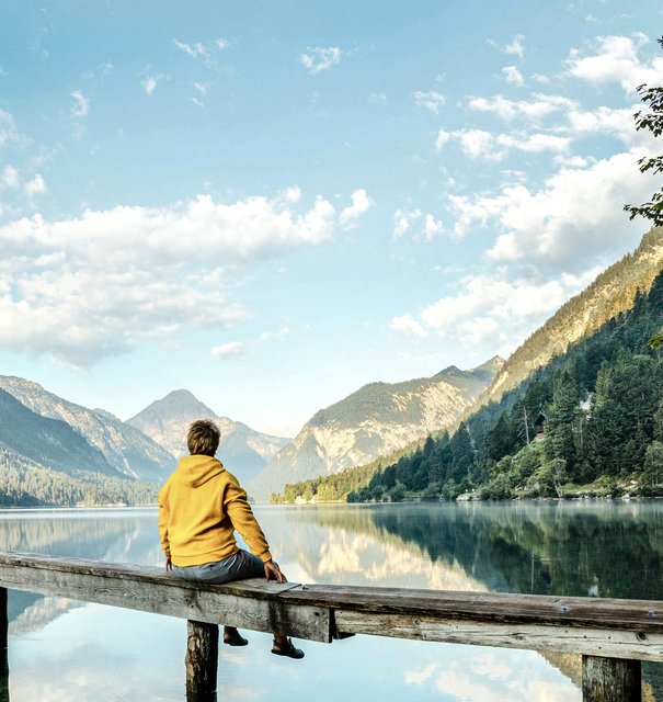 Ein junger Mann in einem gelben Hoodie sitzt auf einem Holzsteg und blickt auf einen ruhigen See, umgeben von Bergen.