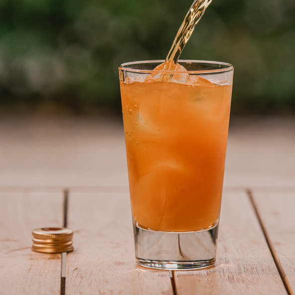 Ein Glas mit einem orangefarbenen Getränk wird auf einem Holztisch gefüllt, während Eiswürfel sichtbar sind.