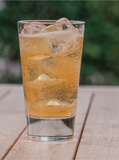 Ein Glas mit einem sprudelnden, goldenen Getränk und Eiswürfeln steht auf einem Holztisch im Freien.