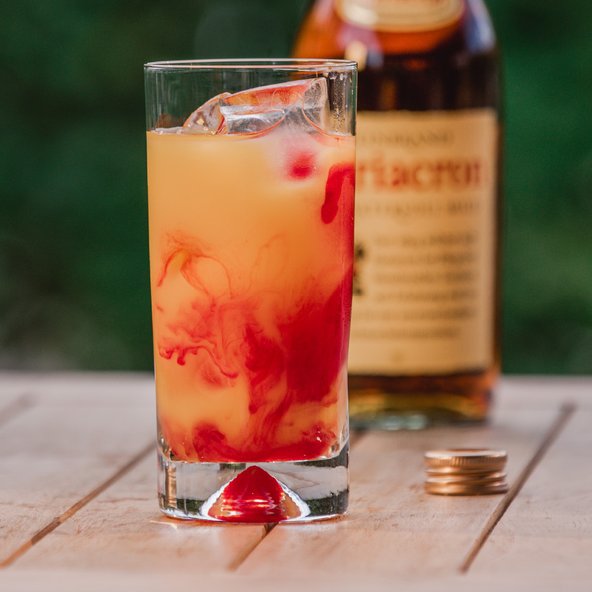 Ein erfrischendes Getränk in einem hohen Glas, mit Eiswürfeln und einer rot-gelben Farbverlauf, auf einem Holztisch.