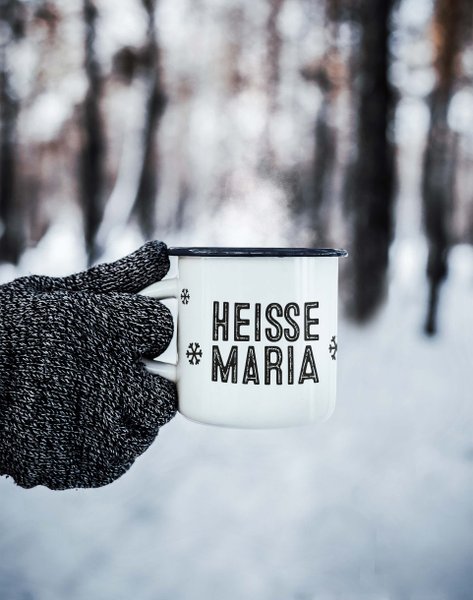 Eine Hand in einem grauen Handschuh hält eine dampfende Tasse mit der Aufschrift 'HEISSE MARIA' vor einer verschneiten Waldlandschaft.