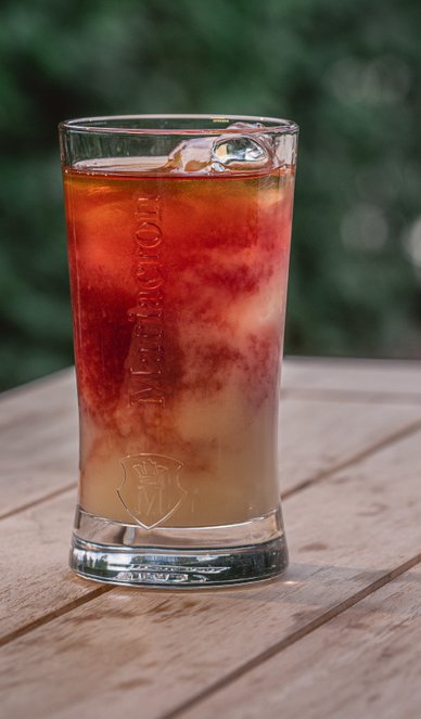 Ein Glas mit einem bunten Getränk, das Eiswürfel enthält, steht auf einem Holztisch vor einem unscharfen grünen Hintergrund.