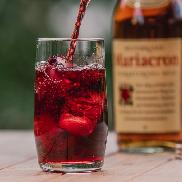 Ein Glas, das mit einem roten, sprudelnden Getränk gefüllt ist, während eine Flasche Mariacron im Hintergrund zu sehen ist.