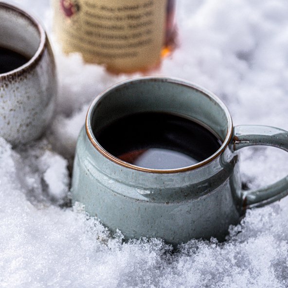 Zwei Tassen mit dampfendem Kaffee stehen in frischem, weißem Schnee, während eine Flasche im Hintergrund sichtbar ist.