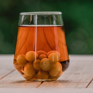 Ein Glas mit einem Getränk, gefüllt mit Eis und Oliven, steht auf einem Holztisch, im Hintergrund eine Flasche Mariacron.