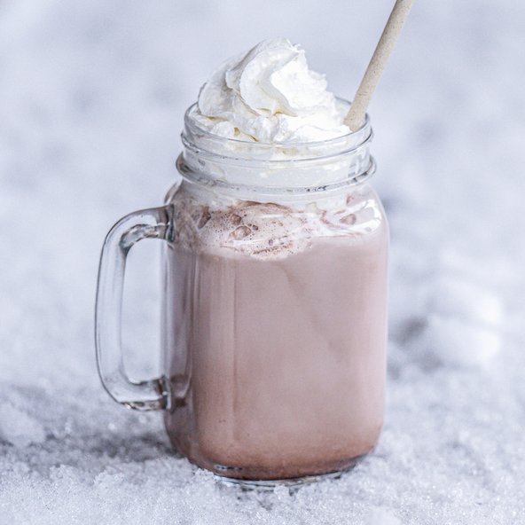 Ein Glas mit heißer Schokolade, gekrönt von einer großzügigen Portion Schlagsahne, steht auf einer schneebedeckten Fläche.
