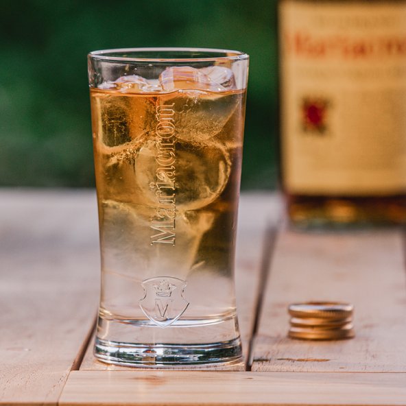 Ein Glas mit einem goldenen Getränk und Eiswürfeln steht auf einem Holztisch, im Hintergrund ist eine Flasche sichtbar.