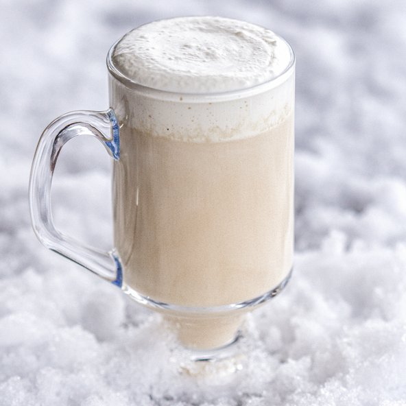 Ein Glas mit schaumigem Getränk steht auf einer schneebedeckten Fläche, das Getränk hat eine cremige, beige Farbe.