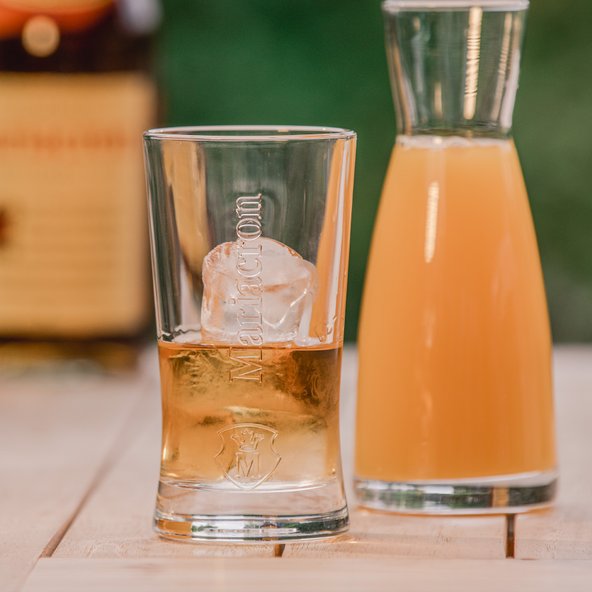 Ein Glas mit einem Eiswürfel und einem goldenen Getränk steht neben einer Karaffe mit orangefarbener Flüssigkeit.