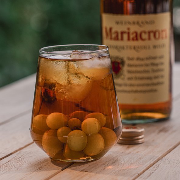 Ein Glas mit einem Getränk, gefüllt mit Eis und Oliven, steht auf einem Holztisch, im Hintergrund eine Flasche Mariacron.