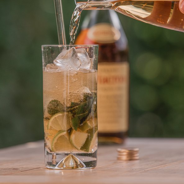Ein Glas mit Eis, Limettenscheiben und frischer Minze wird mit einem sprudelnden Getränk gefüllt, während eine Flasche im Hintergrund sichtbar ist.