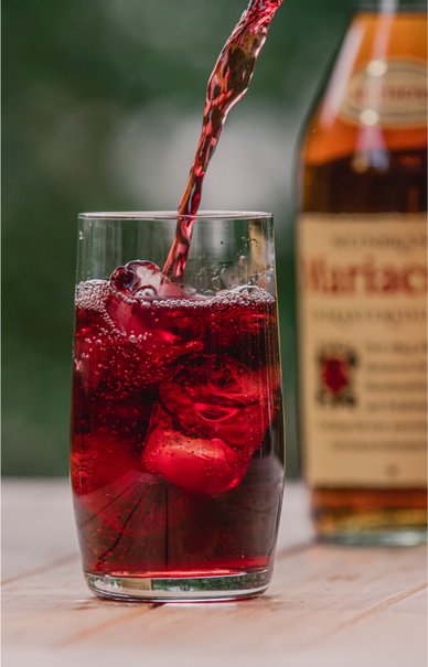 Ein Glas, das mit einem roten, sprudelnden Getränk gefüllt ist, während eine Flasche Mariacron im Hintergrund zu sehen ist.