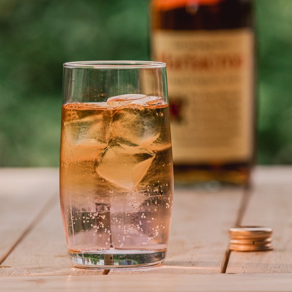 Ein Glas mit sprudelndem Getränk und Eiswürfeln steht auf einem Holztisch, im Hintergrund eine Flasche mit Etikett.