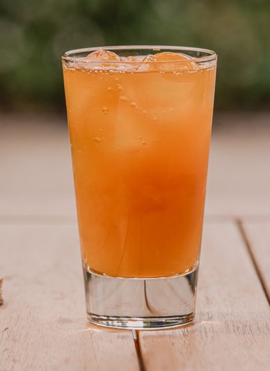 Ein Glas mit einem orangefarbenen Getränk wird auf einem Holztisch gefüllt, während Eiswürfel sichtbar sind.