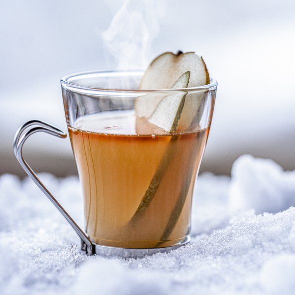 Ein dampfender Tee in einem klaren Glas, garniert mit einer Scheibe Birne, steht auf frischem, weißem Schnee.