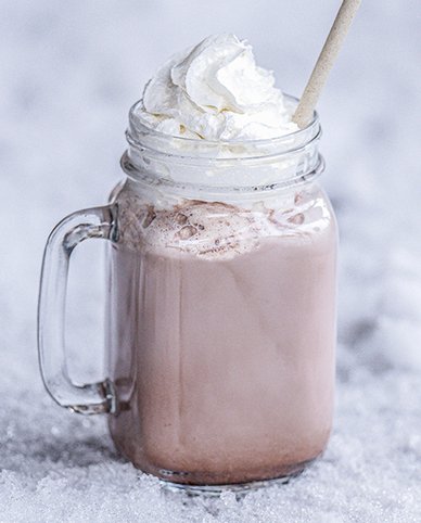 Ein Glas mit heißer Schokolade, gekrönt von einer großzügigen Portion Schlagsahne, steht auf einer schneebedeckten Fläche.